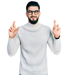 Young arab man with beard wearing elegant turtleneck sweater and glasses gesturing finger crossed smiling with hope and eyes closed. luck and superstitious concept.