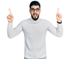 Young arab man with beard wearing elegant turtleneck sweater and glasses smiling amazed and surprised and pointing up with fingers and raised arms.