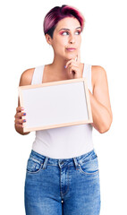 Young beautiful woman with pink hair holding empty white chalkboard serious face thinking about question with hand on chin, thoughtful about confusing idea