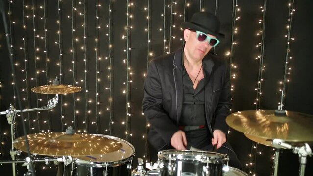 Man in glasses and shirt plays drum set on studio with garlands.