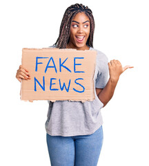 Young african american woman with braids holding fake news banner pointing thumb up to the side...