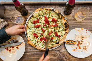 Male hand taking a slice of freshly baked Neapolitan zucchini pizza on a rustic wooden table in a traditional Pizzeria. - 710929155