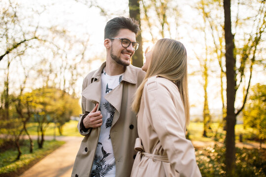 Portrait of a guy and a woman posing and smiling with teeth laughing in beige trench coats in the park at sunset. Spring sunny landscape. Concept of beauty, love and happiness.