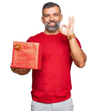 Middle age handsome man holding gift doing ok sign with fingers, smiling friendly gesturing excellent symbol