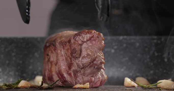 Close-up chef flips juicy steak roasting with rosemary and garlic on a frying pan on induction stove. Steam rising above the frying pan. Cooking at home. Slow motion