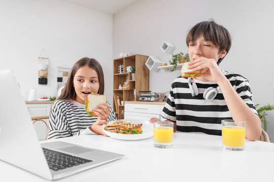 Cute children with laptop, tasty sandwiches and glasses of juice watching movie in kitchen