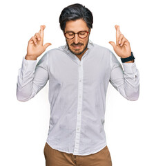 Naklejka premium Young hispanic man wearing business shirt and glasses gesturing finger crossed smiling with hope and eyes closed. luck and superstitious concept.