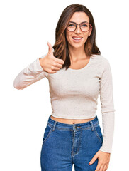 Young brunette woman wearing casual clothes and glasses doing happy thumbs up gesture with hand. approving expression looking at the camera showing success.