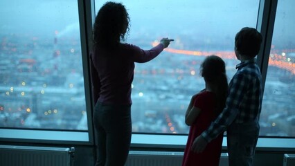 Mother shows children evening cityscape in large window