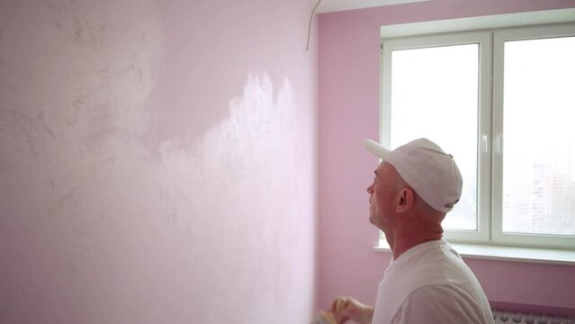 Builder in white cap is painting wall at the light pink room.