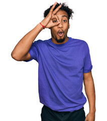 Young african american man with beard wearing casual purple t shirt doing ok gesture shocked with surprised face, eye looking through fingers. unbelieving expression.