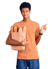 Young african american man holding paper bag with bread pointing aside worried and nervous with...