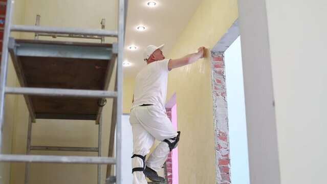 Finisher is standing on stepladder and polishing wall near ceiling