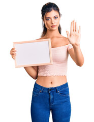 Young beautiful woman holding empty white chalkboard with open hand doing stop sign with serious and confident expression, defense gesture