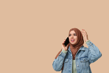 Beautiful young Muslim woman in hijab talking by mobile phone on beige background