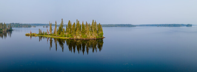 An aerial panoramic view of a large calm lake with a small island that is covered in a forest of spruce and pine trees. The calm water reflects the trees and the blue sky.
 - Powered by Adobe