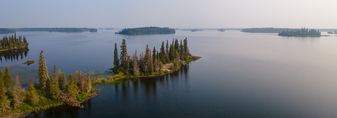 A panoramic aerial view of a large northern lake with a prominent rocky point of land that is covered in spruce and pine trees.  The calm water reflects the color of the pale blue sky.
