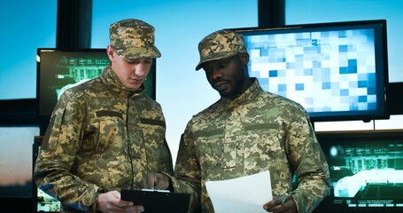 Mixed-races male soldiers in uniforms talking in office with screens and computers. Caucasian and African American militarians reading documents and shaking hands when saying goodbye.