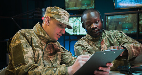 Two mixed-races army officers talking about secret operation. African American military dispatcher sitting in office and Caucasian male soldier bringing documents with new data and information.