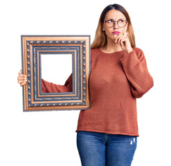 Beautiful young woman holding empty frame serious face thinking about question with hand on chin,...