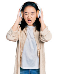 Young chinese woman listening to music using headphones angry and mad screaming frustrated and furious, shouting with anger. rage and aggressive concept.