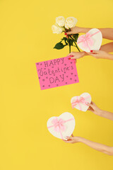 Female hands holding sign with text HAPPY VALENTINE'S DAY, heart-shaped gift boxes and roses on yellow background
