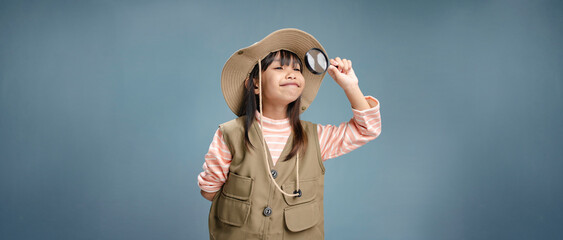 A little girl is wearing a camping clothes, holding a magnifying glass for studying.