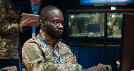 Portrait shot of African American young man in glasses sitting at laptop computer and typing on keyboard, then looking at camera in monitoring room in army. Soldier working in military data center.