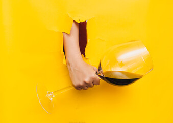 A man's hand emerges through a torn hole in yellow paper with a large glass of fresh red wine. The...