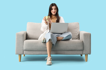 Fototapeta na wymiar Beautiful young woman with cup of tea and laptop on sofa against blue background