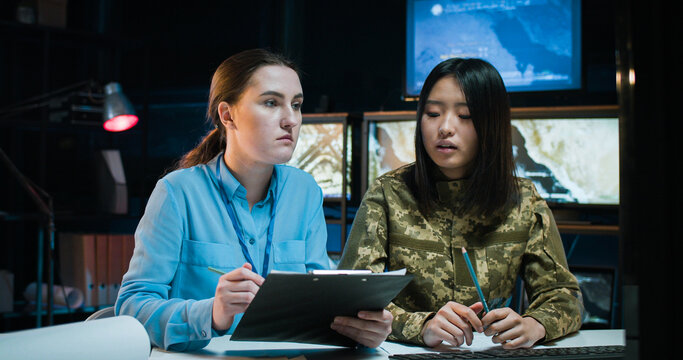 Female Caucasian office worker and soldier sitting together at desk in monitoring center with screens and studying some militarian documents. Women talking and discussing army strategy.