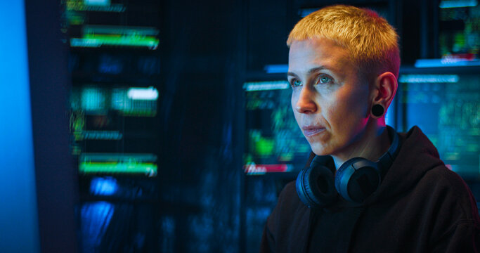 Portrait of Caucasian woman hacker with blond short hair sitting in dark room with monitors at desk in front of computer. Turning face to camera and taking on hood. Close up of female hacking system.