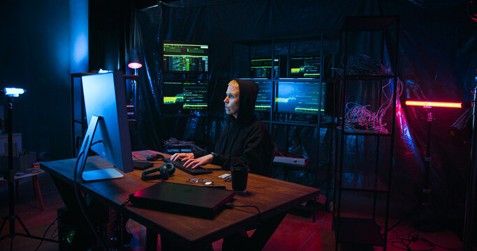Anonymous Caucasian female hacker typing on keyboard and breaking secure system of computer in dark room with monitors. Woman with masked face commiting cybercrime online and stealing information.