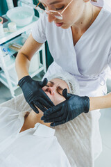 A dermatologist treats the face of a woman lying on a massage table. Facial skin care. A female beautician applies a mask to a woman's face