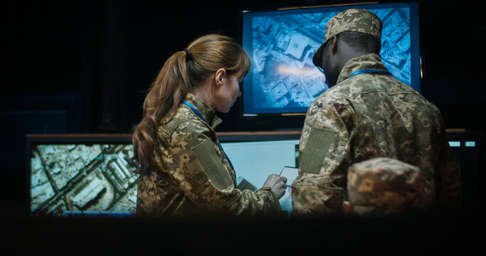 African American man general in camouflage uniform standing in monitoring room and watching troops dislocation on screens. Caucasian woman coming and showing some documents with daa and info.