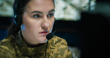 Caucasian military woman in headset sitting at computer screen and talking via videochat in...