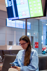 Beautiful young woman looking on smartphone checking the flight schedule while waiting boarding on aircraft in airport lounge