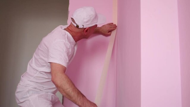 Painter in white clothes and kneepads sticks masking tape