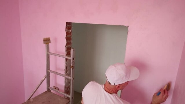 Worker in cap and white clothes paints wall in room