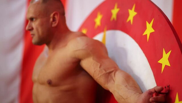 Bodybuilder stands his back to disk-target for throwing knives 