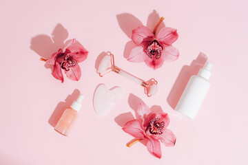 Facial cosmetic roller and gua sha scraper with pink orchid flowers and serum bottles. Sharp shadows. Home spa concept. Top view, flat lay
