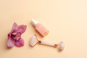 Pink serum bottle, rose quartz crystal facial roller and orchid flower on beige background. Top view, flat lay, copy space