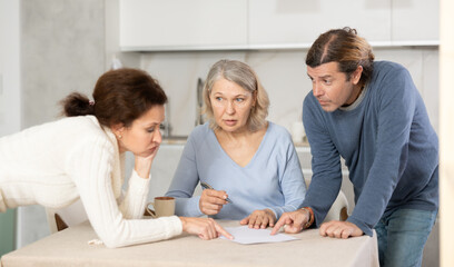 Elderly woman sits in kitchen and signs contract in presence of adult children. Documenting intentions and actions.