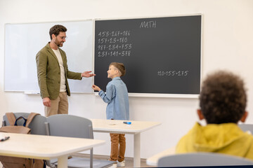 Little boy standing in classroom while learning math lesson with his teacher.