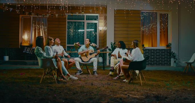 Joyful different people resting in backyard in summer night, playing guitar and singing, 4K, Prores