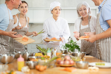 Friendly skilled young Asian female chef giving culinary classes to group of men and women of...