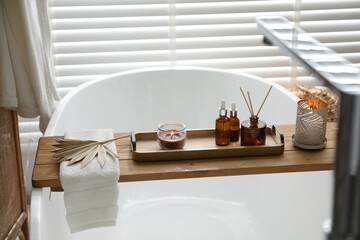 Wooden tray with cosmetic products, burning candles, reed air freshener and towel on bath tub in...