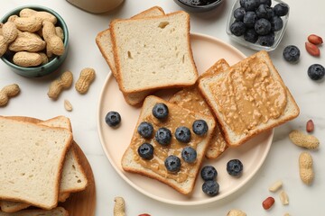 Delicious toasts with peanut butter, blueberries and nuts on white marble table, flat lay