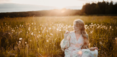 young blonde woman sitting in a field with dandelions in the summer at sunset. Summer holiday...