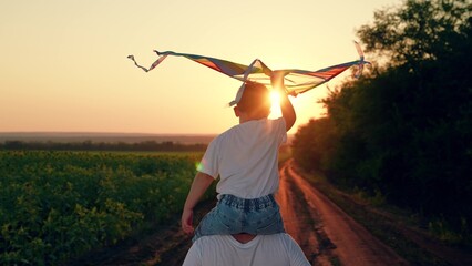 Happy boy on dad shoulders runs along country road, plays with toy kite, sunset. Child, dad is...